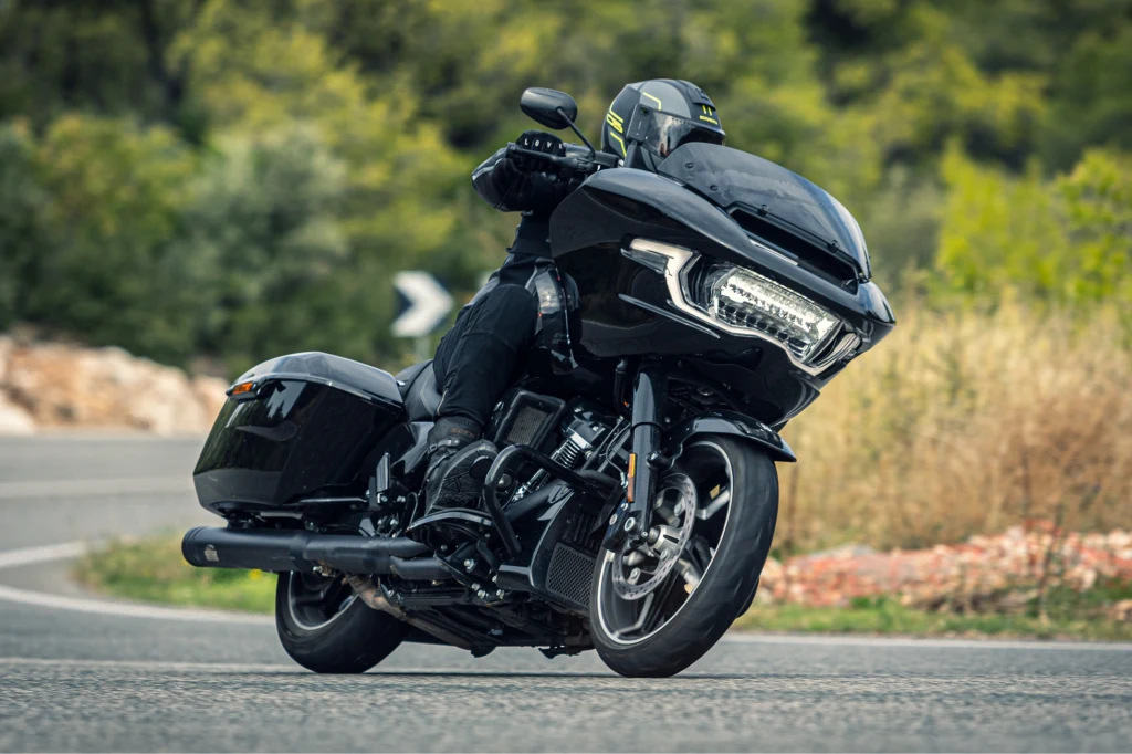 Motocykl turystyczny o masywnej sylwetce skręcający na zakręcie, kierowca w czarnym kombinezonie i kasku, otoczony zielenią i asfaltową drogą.