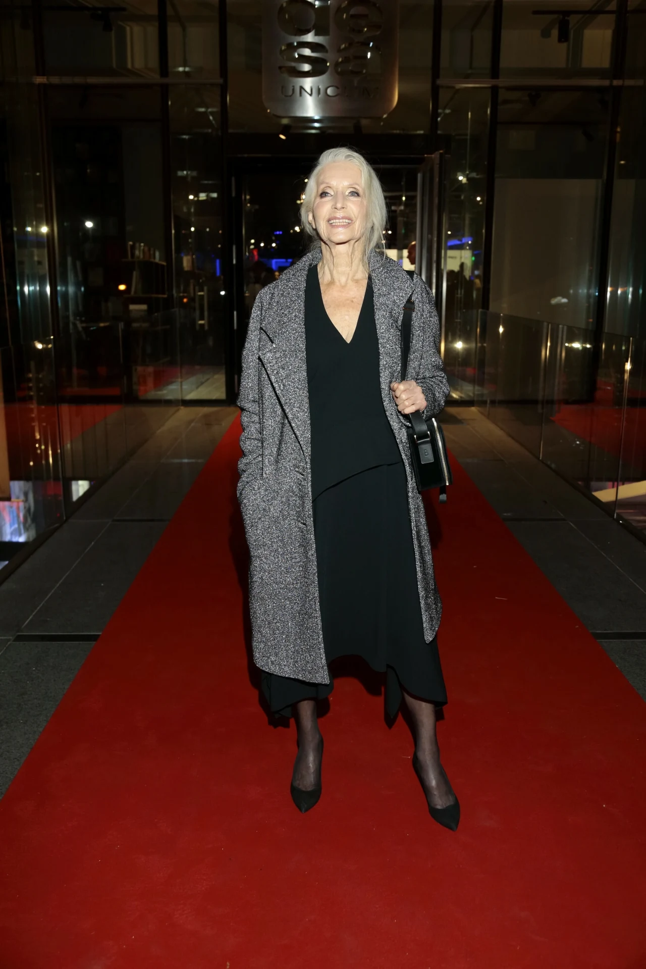 Helena Norowicz w szarym płaszczu do połowy łydki Elegancka starsza kobieta w szarym płaszczu i czarnej sukni stoi na czerwonym dywanie przed wejściem do budynku, trzymając czarną torebkę i uśmiechając się lekko.