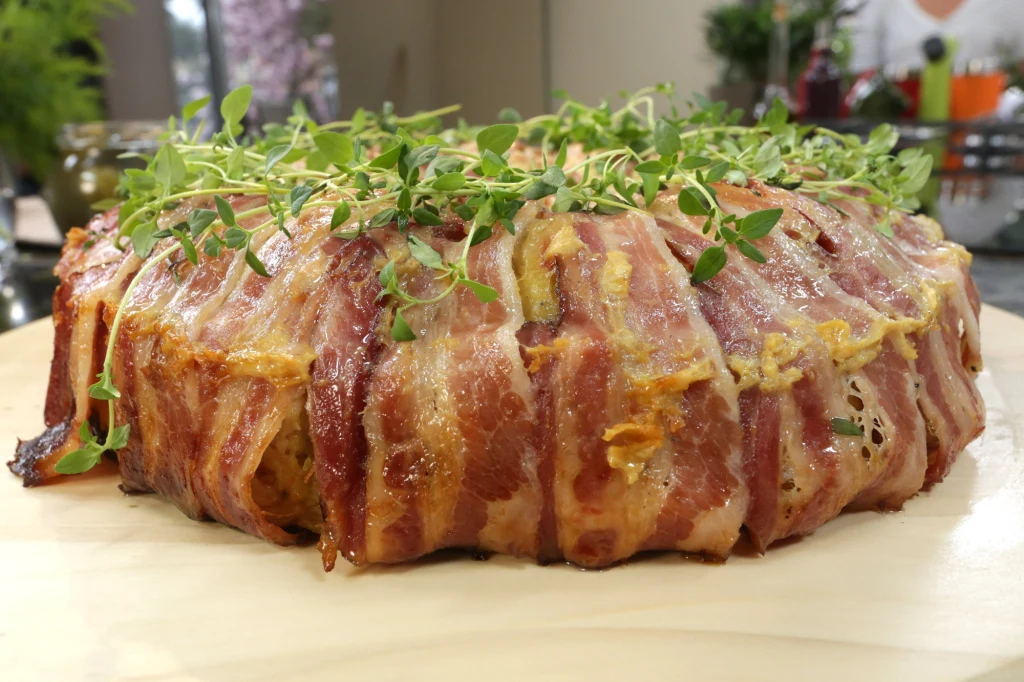 Pieczona potrawa owinięta plastry bekonu, udekorowana świeżymi ziołami, ułożona na drewnianej desce.