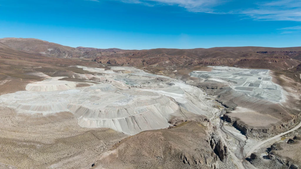 Wydobycie litu w Argentynie Rozległa kopalnia odkrywkowa rozciągająca się na pustynnym, górzystym terenie z organizacją wyrobisk i dróg technicznych, otoczona pagórkami i wzniesieniami pod błękitnym niebem.