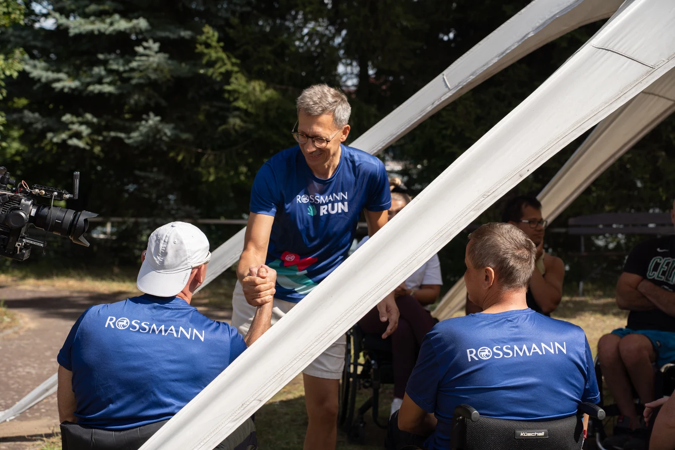 Rossmann Grupa mężczyzn ubranych w sportowe koszulki z napisem Rossmann Run podczas wydarzenia plenerowego, jeden z nich uśmiecha się i ściska dłoń innemu mężczyźnie siedzącemu na wózku inwalidzkim, całość odbywa się pod namiotem.