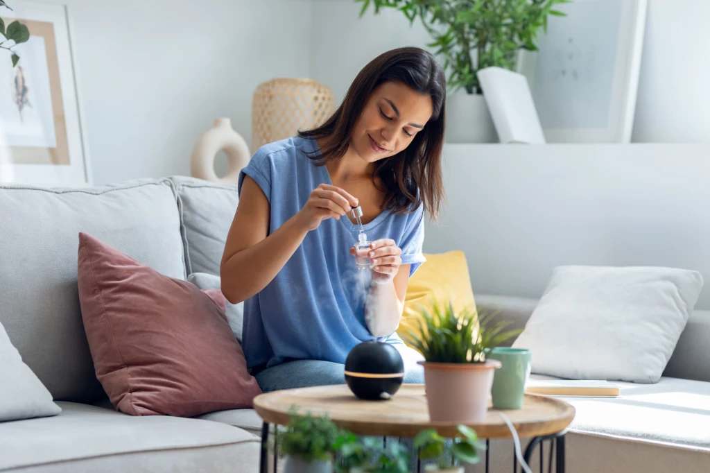 Prosta zmiana w domu, która od razu odmładza skórę Kobieta siedzi na jasnej sofie w przytulnym salonie, przygotowując aromaterapeutyczny dyfuzor z olejkami eterycznymi. Na stoliku stoją rośliny doniczkowe, notes i ciemna kulista lampka. W tle widoczne są dekoracje i jasne ściany.