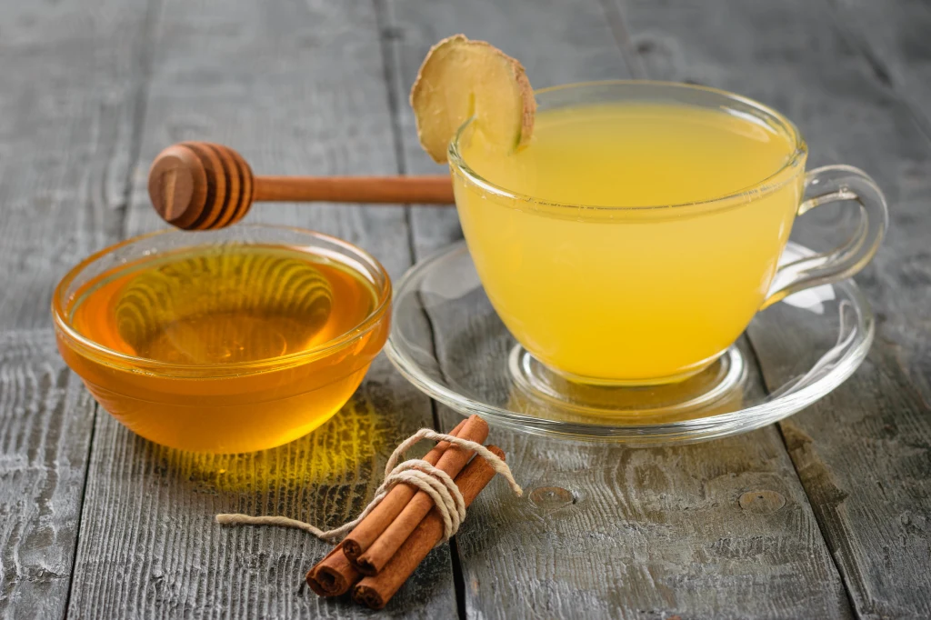 Napój miodowo-cynamonowy - przepis Ewy Wachowicz Szklanka gorącego napoju z plastrem imbiru na krawędzi, miseczka miodu z drewnianą łyżeczką oraz pałeczki cynamonu przewiązane sznurkiem na drewnianym stole.