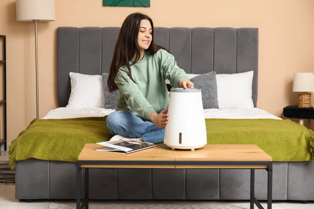 Sucha cera i widoczne linie? To, czym oddychasz, może być największym wrogiem skóry Młoda kobieta siedzi na łóżku w nowocześnie urządzonej sypialni i ustawia biały nawilżacz powietrza stojący na stoliku. W tle szare wezgłowie łóżka oraz zielona narzuta. Obok na stoliku leży otwarty magazyn.