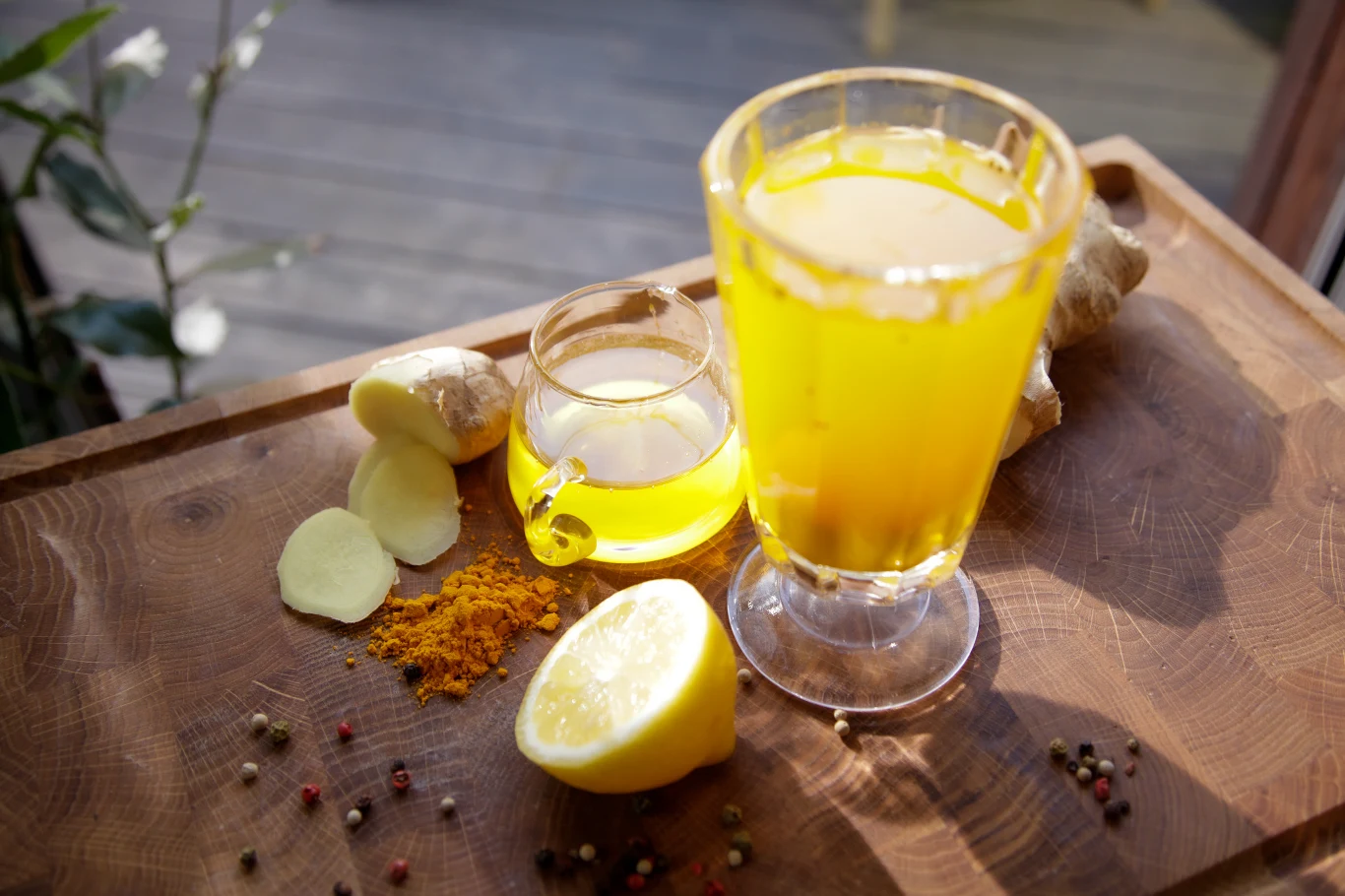 Napój z imbirem i kurkumą - przepis Ewy Wachowicz Szklanka żółtego napoju ustawiona na drewnianej desce, obok plasterki imbiru, kawałek cytryny, sproszkowana kurkuma, ziarna pieprzu oraz niewielki dzbanuszek z żółtym płynem.