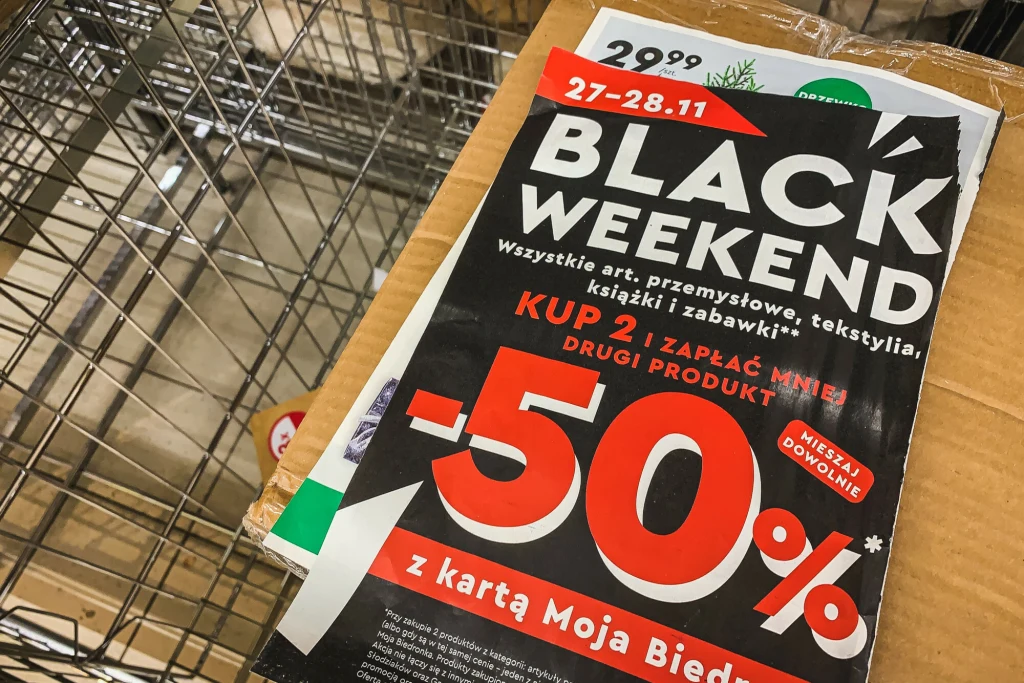 Ulotka reklamowa informująca o promocji Black Weekend w sklepie, oferująca 50% zniżki na drugi produkt przy zakupie dwóch artykułów przemysłowych, tekstyliów, książek i zabawek, widoczna na tle metalowego koszyka sklepowego.