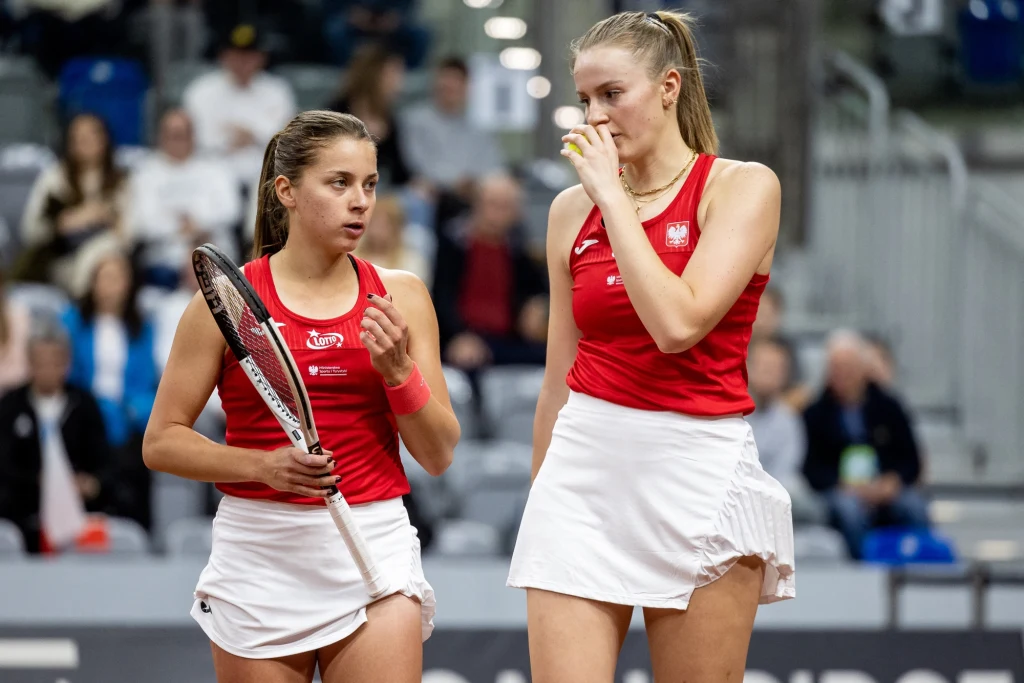 Martyna Kubka (z prawej), obok Maja Chwalińska Dwie tenisistki w czerwonych koszulkach i białych spódniczkach dyskutują podczas meczu, jedna z nich trzyma rakietę tenisową, w tle rozmazana publiczność na trybunach.