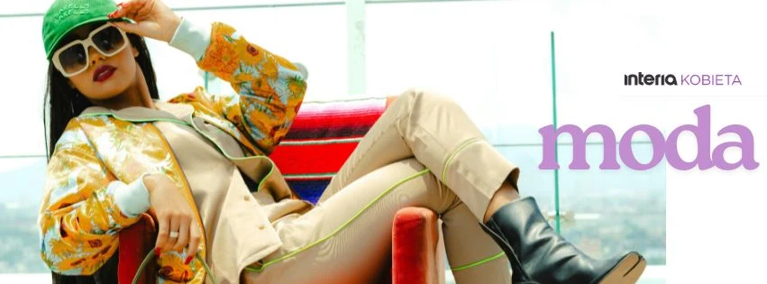 Stylowe inspiracje, które odmienią każdy look Stylowa kobieta w modnym ubraniu, z zieloną czapką, okularami przeciwsłonecznymi, kolorową kurtką i jasnymi spodniami, siedzi wygodnie na czerwonym fotelu na tle przeszklonego tarasu.