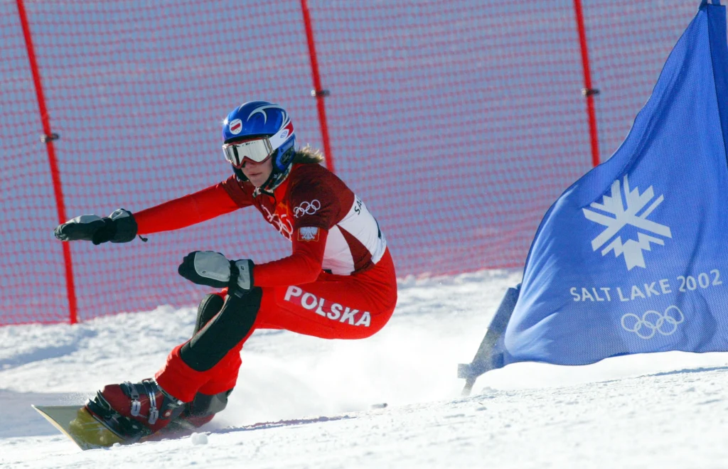 Salt Lake City 2002. Jagna Marczułajtis jeszcze w czasach kariery zawodniczej Zawodniczka reprezentująca Polskę w czerwonym stroju olimpijskim zjeżdża na snowboardzie po śnieżnym stoku, mijając niebieską flagę z napisem Salt Lake 2002 oraz logo zimowych igrzysk olimpijskich, w tle siatka zabezpieczająca