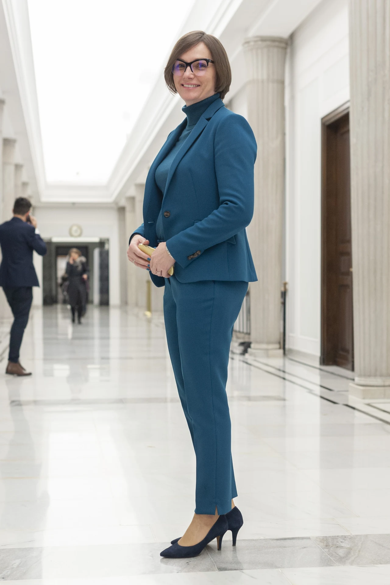 Jagna Marczułajtis-Wlaczak jako parlamentarzystka Kobieta w okularach ubrana w niebieski garnitur i czarne szpilki uśmiecha się stojąc w jasnym, eleganckim korytarzu z białymi ścianami i kolumnami. W tle widoczne są inne osoby.