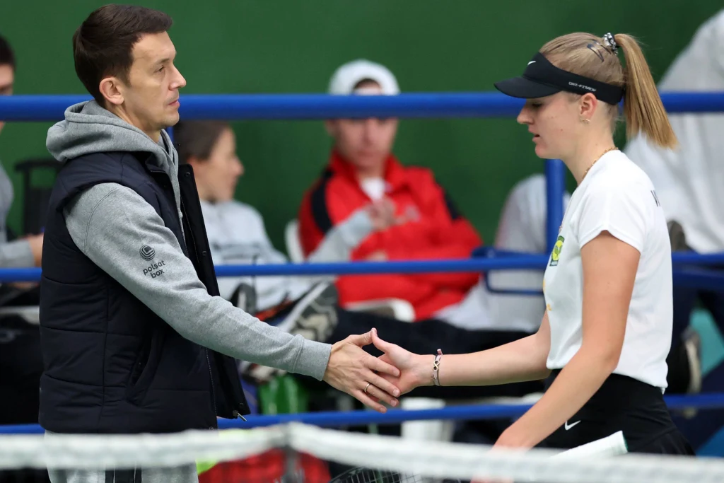 Dwie osoby podają sobie dłonie na tle trybun, kobieta ubrana w sportowy strój i czapkę z daszkiem, mężczyzna w czarnej kamizelce i szarej bluzie. W tle widoczne rozmazane postacie kibiców.