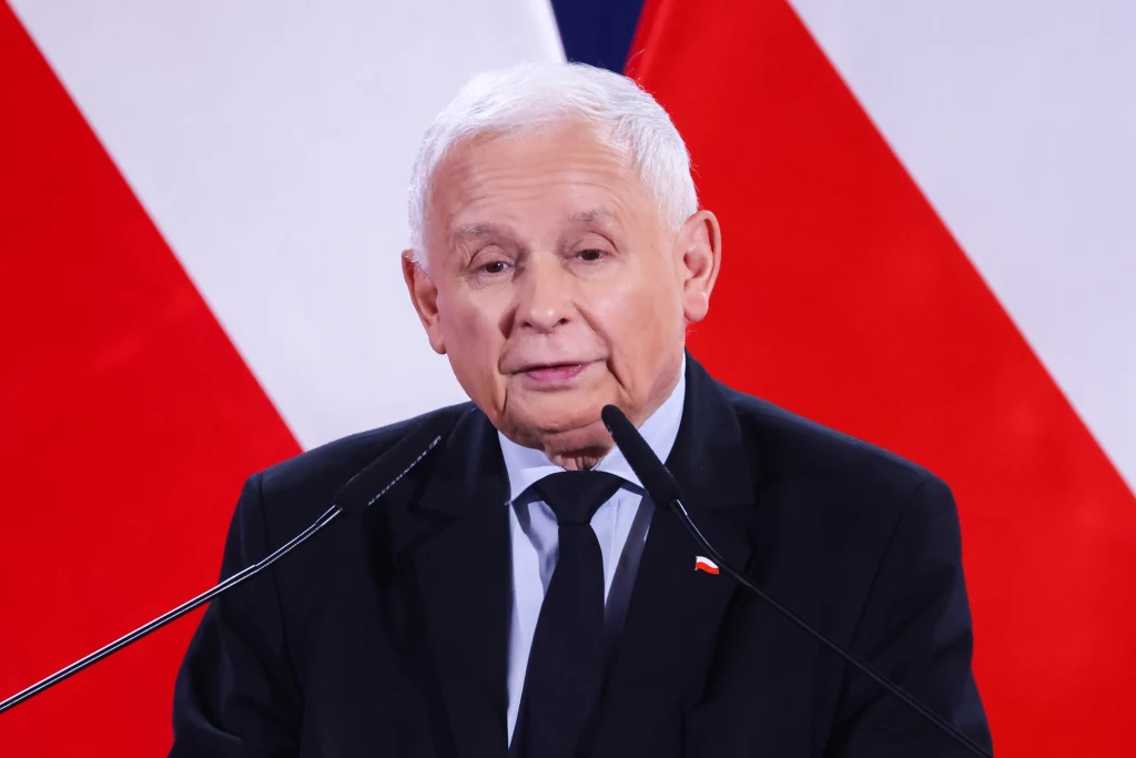 Starszy mężczyzna w garniturze podczas wystąpienia publicznego. W tle znajdują się biało-czerwone flagi. Przemawia do mikrofonów, prezentuje poważny wyraz twarzy.