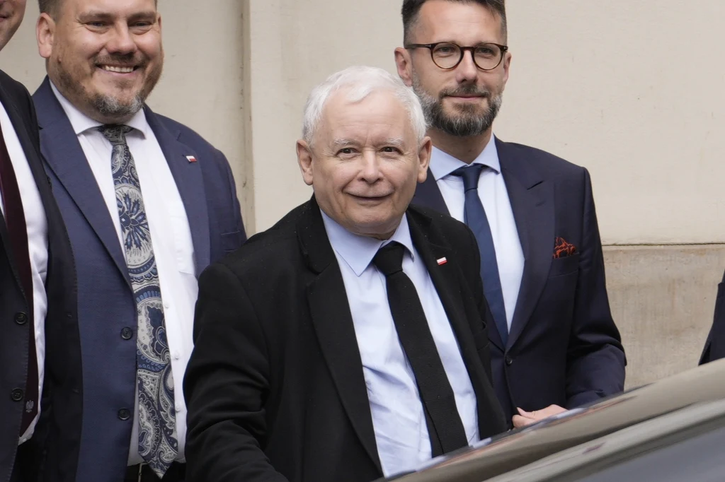 Trzech mężczyzn w garniturach stoi blisko siebie, jeden z nich jest starszy, ma białe włosy i uśmiecha się lekko w stronę aparatu, pozostali również mają pozytywne wyrazy twarzy, tło stanowi jasna ściana budynku.