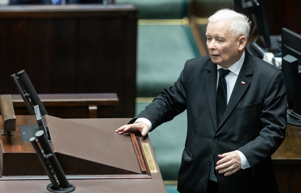 Starszy mężczyzna w ciemnym garniturze stoi przy mównicy sejmowej w sali posiedzeń parlamentu. Na jego klapie widoczna jest przypinka w barwach narodowych. W tle widać fragmenty ław sejmowych.