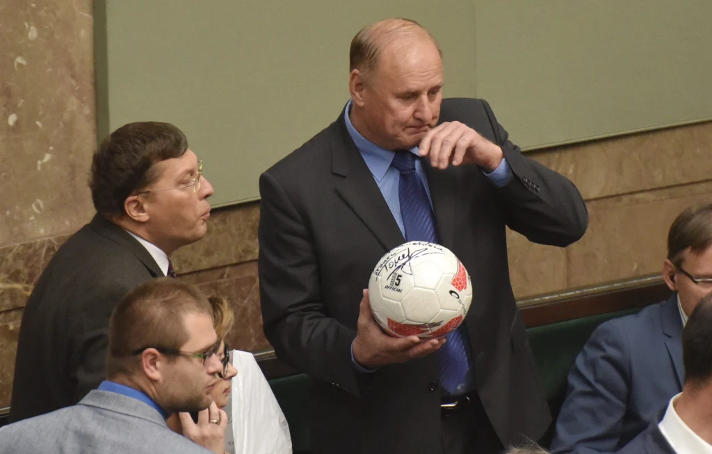 Mężczyzna w garniturze trzyma podpisaną piłkę nożną, rozmawia z innym mężczyzną otoczonym przez grupę osób w formalnych strojach, w tle widoczne ławki przypominające salę obrad.
