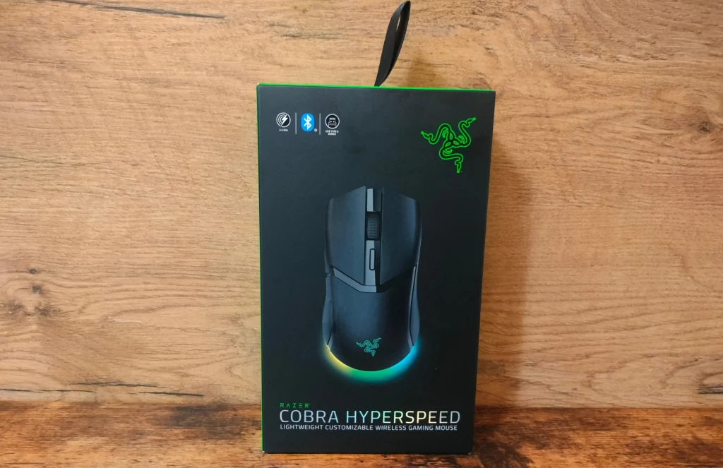 Pudełko myszki Razer Cobra Hyperspeed z widocznym logo firmy, grafiką przedstawiającą mysz oraz informacjami o funkcjach na tle drewnianej powierzchni.