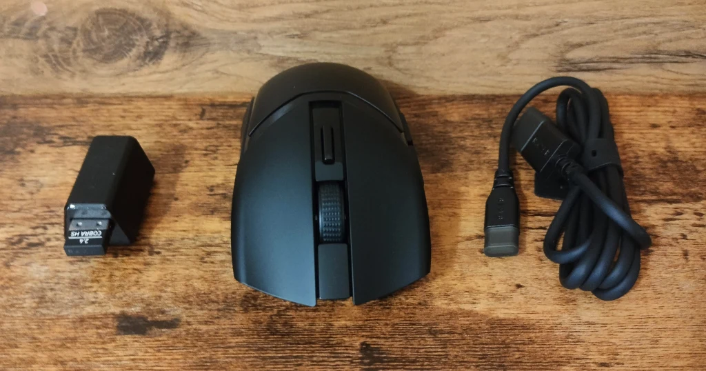 Bezprzewodowa mysz komputerowa w kolorze czarnym wraz z odbiornikiem USB oraz kablem zasilającym typu USB, ułożone na drewnianym blacie.