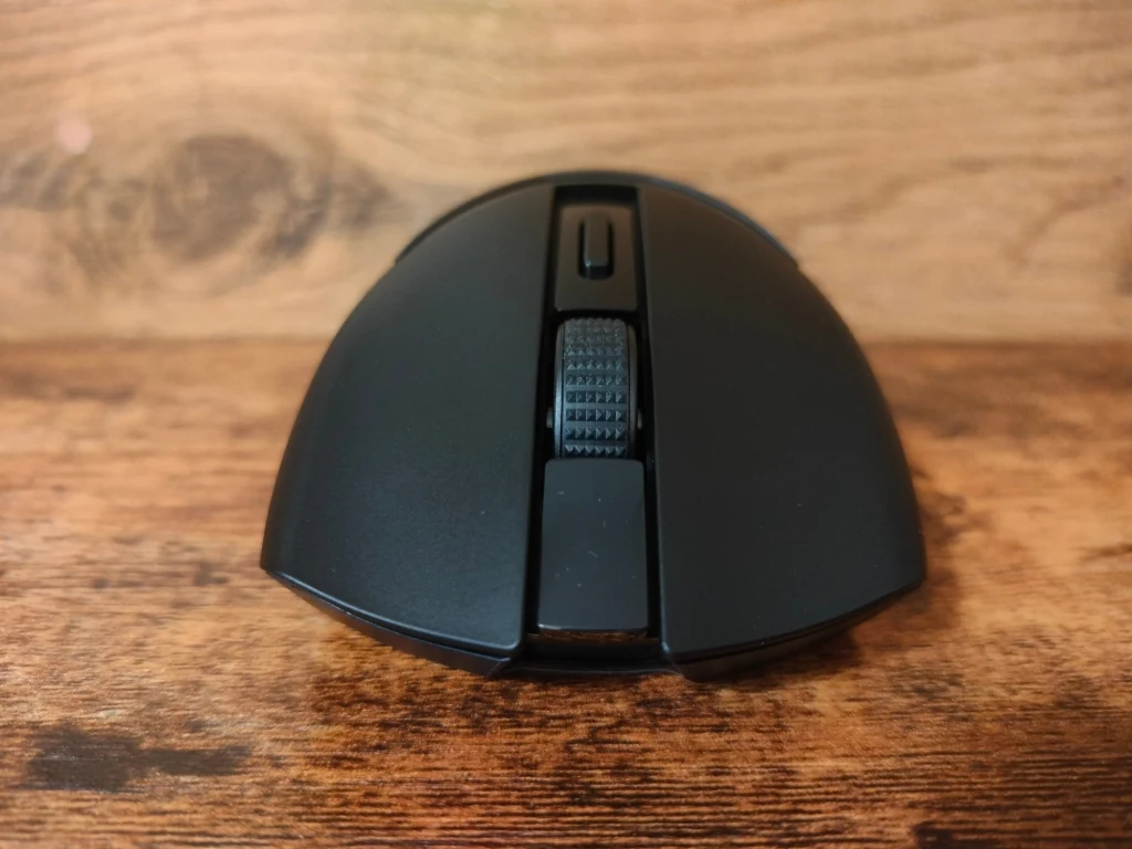 Czarna bezprzewodowa mysz komputerowa na drewnianym tle, widok z przodu. Wyraźnie widoczna rolka przewijania i przyciski sterujące.