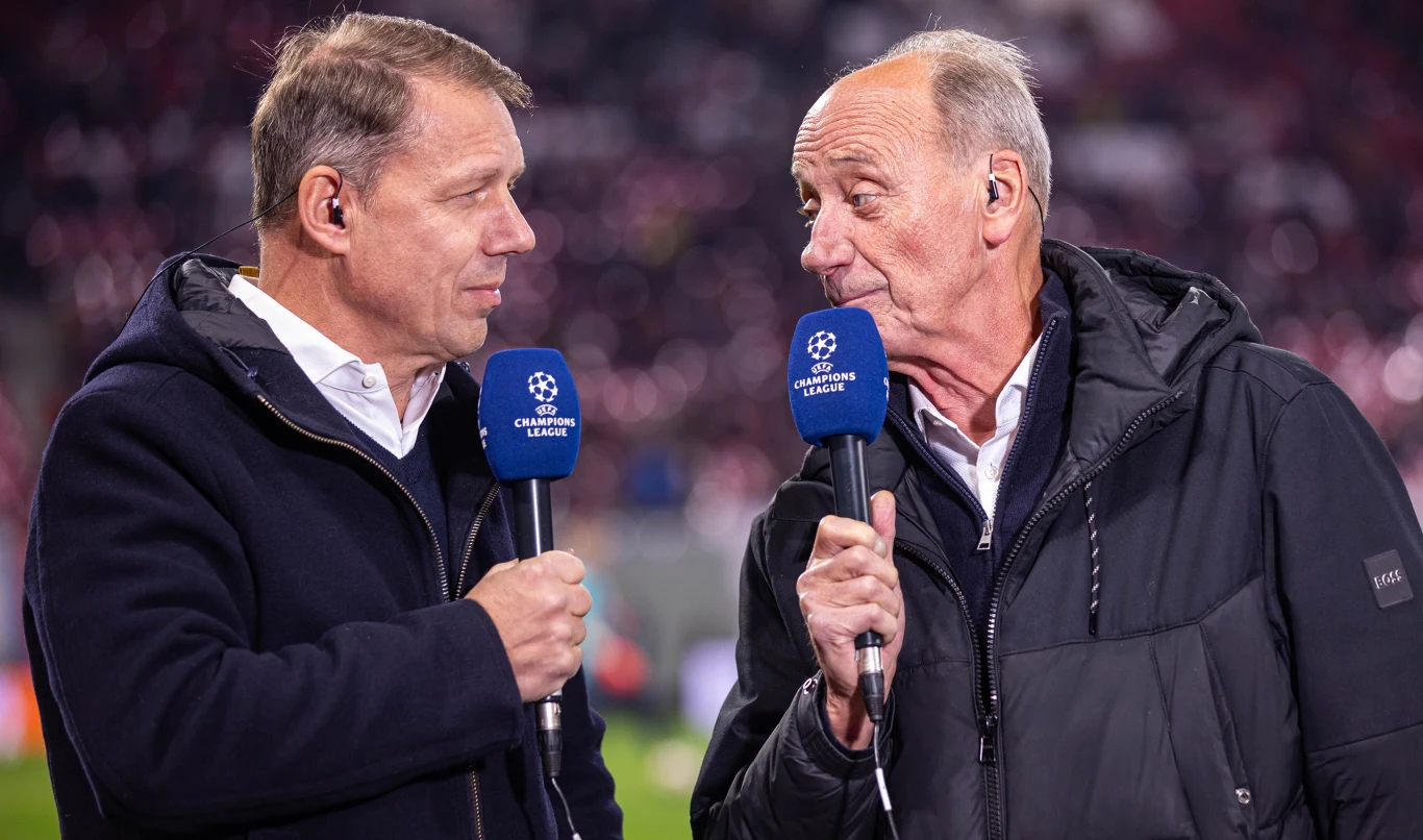 Dwóch mężczyzn rozmawiających ze sobą podczas relacji telewizyjnej, trzymają w rękach mikrofony z logo Ligi Mistrzów UEFA, ubrani w ciemne kurtki, w tle rozmyte światła stadionu.