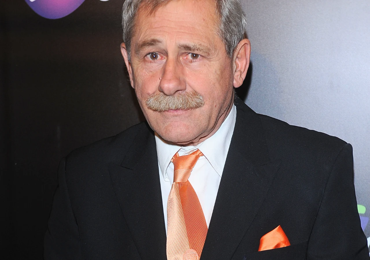 Andrzej Strzelecki Starszy mężczyzna w garniturze z kremową koszulą oraz pomarańczowym krawatem i poszetką stoi na tle ciemnej ścianki z kolorowymi elementami graficznymi.