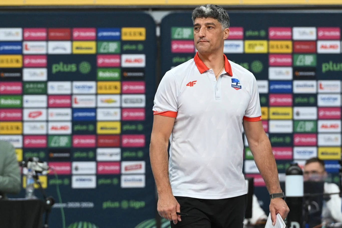 Trener siatkówki w białej koszulce z logotypem drużyny stoi na tle wielobarwnej ściany pełnej logotypów sponsorów, w tle widać część obsługi technicznej spotkania.