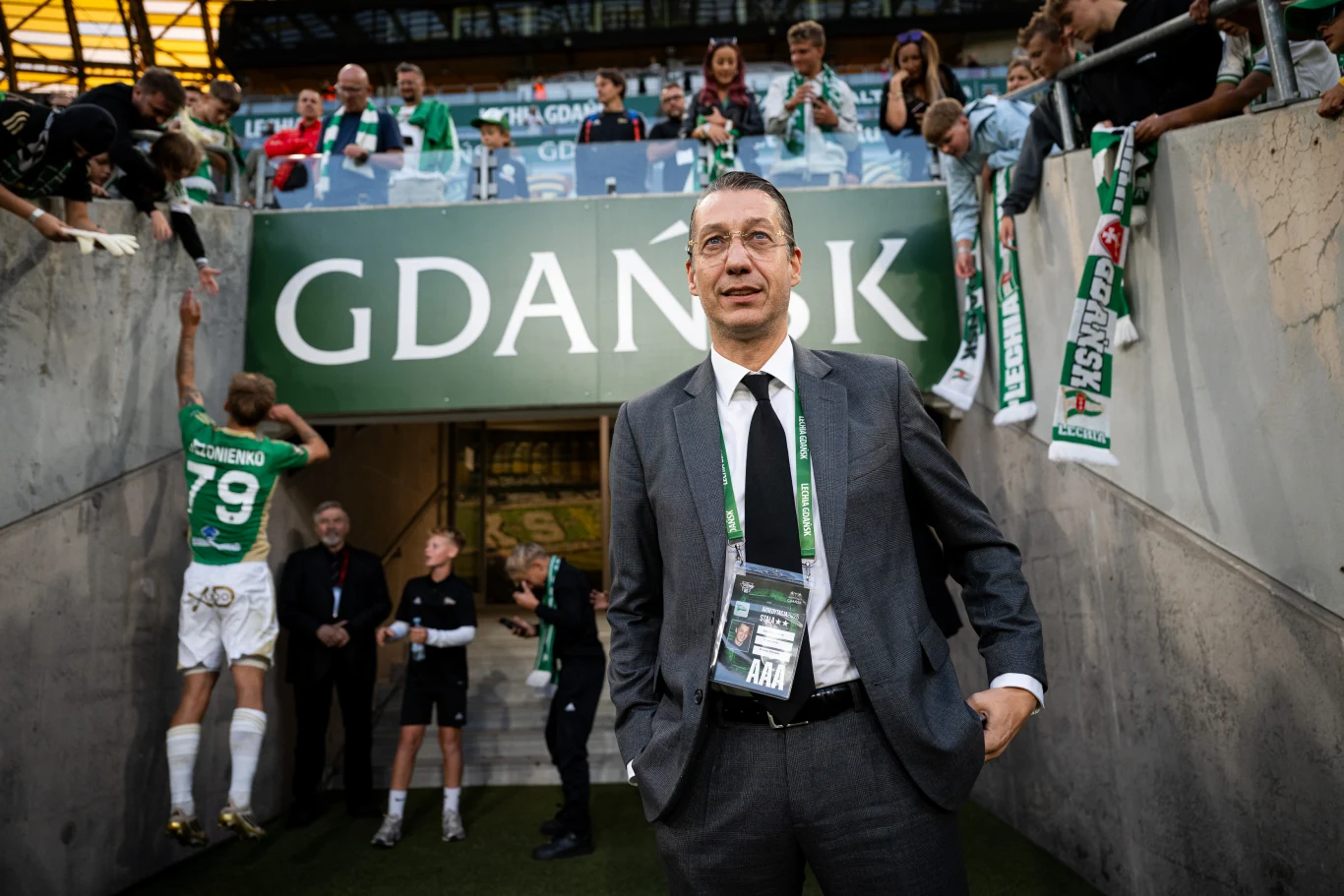 Mężczyzna w garniturze z identyfikatorem na szyi stoi na tle wejścia na stadion z napisem Gdańsk, wokół kibice oraz piłkarze, atmosfera sportowa.