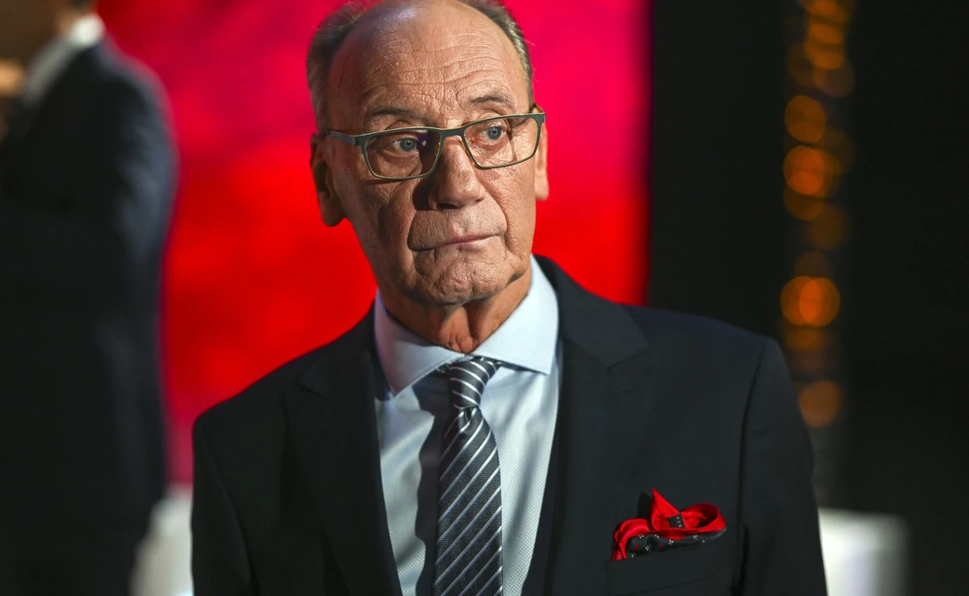 Starszy mężczyzna w eleganckim garniturze z krawatem i kolorową poszetką, w okularach, o poważnym wyrazie twarzy, na tle rozmytego, czerwonego światła.