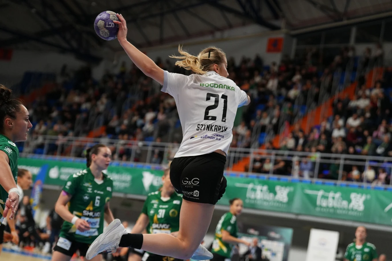 Zawodniczka drużyny piłki ręcznej w białej koszulce z numerem 21 wykonuje dynamiczny skok z piłką do bramki, wokół niej zawodniczki rywalizującej drużyny ubrane na zielono oraz widownia na trybunach.