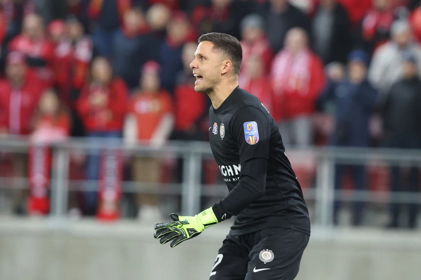 Bramkarz w czarnym stroju sportowym, w jasnozielonych rękawicach, stoi na stadionie piłkarskim podczas meczu, wokół kibice w czerwonych barwach na trybunach.