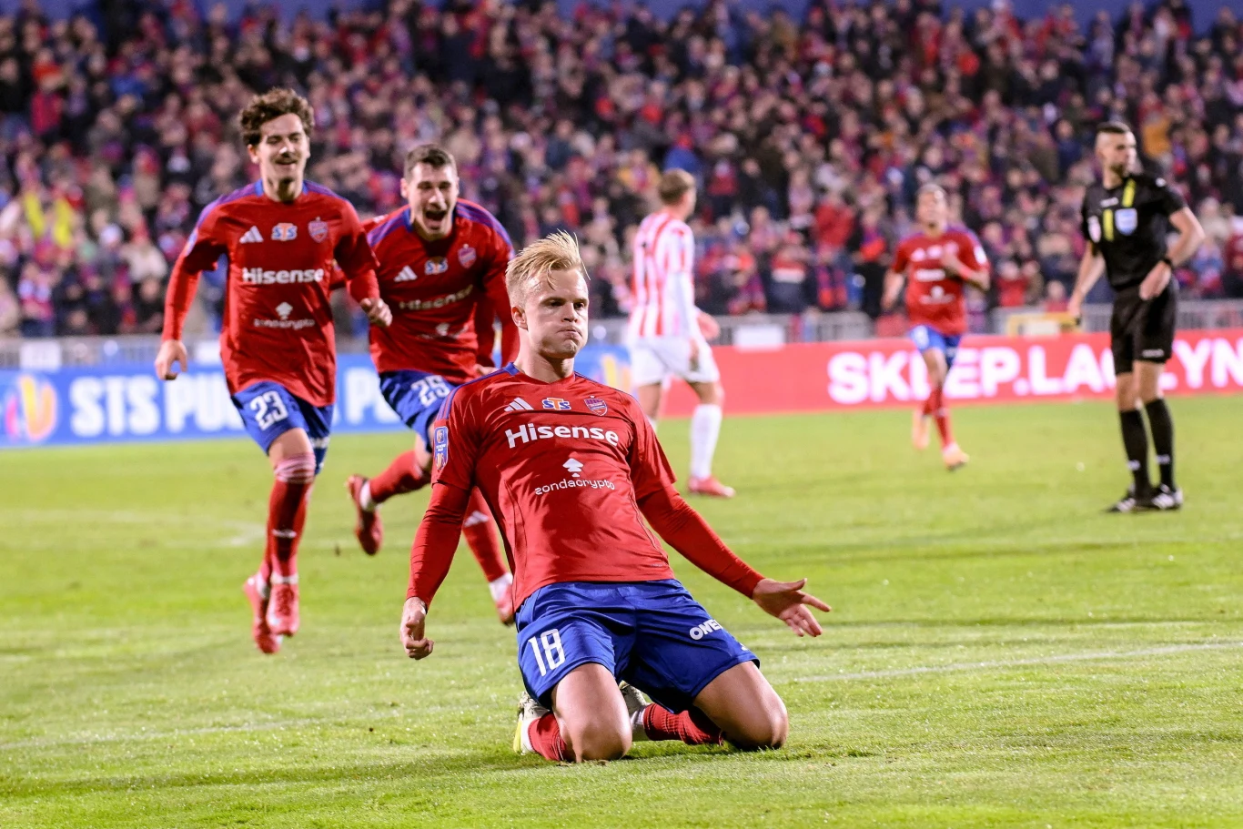 Piłkarz w czerwonym stroju świętuje bramkę, ślizgając się na kolanach po murawie, podczas gdy za nim biegną jego współtowarzysze z drużyny, a w tle widać rozemocjonowaną publiczność na trybunach.