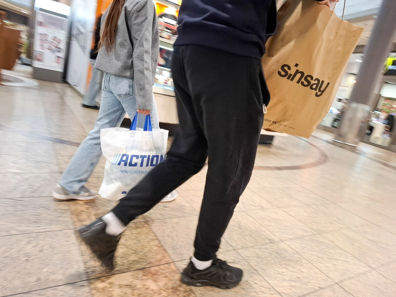 Sinsay wydało komunikat ws. wycofanych produktów Dwie osoby idące przez galerię handlową z torbami zakupowymi z logo sklepów Sinsay i Action, widoczne są fragmenty ich sylwetek oraz wnętrze centrum handlowego.