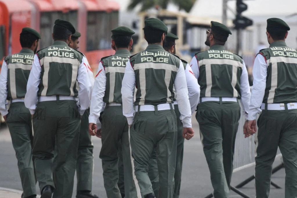 Policja w Dubaju (zdj. ilustracyjne) Grupa policjantów ubranych w mundury z napisem 'POLICE' na plecach idzie ulicą miasta, widoczne są służbowe nakrycia głowy, elementy infrastruktury miejskiej oraz fragmenty czerwonych pojazdów w tle.