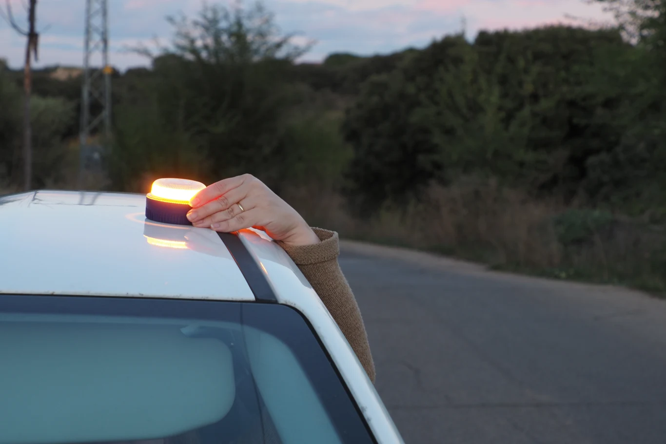 Ręka osoby umieszcza lampę ostrzegawczą na dachu białego samochodu stojącego na poboczu wiejskiej drogi, w tle drzewa i krzewy.