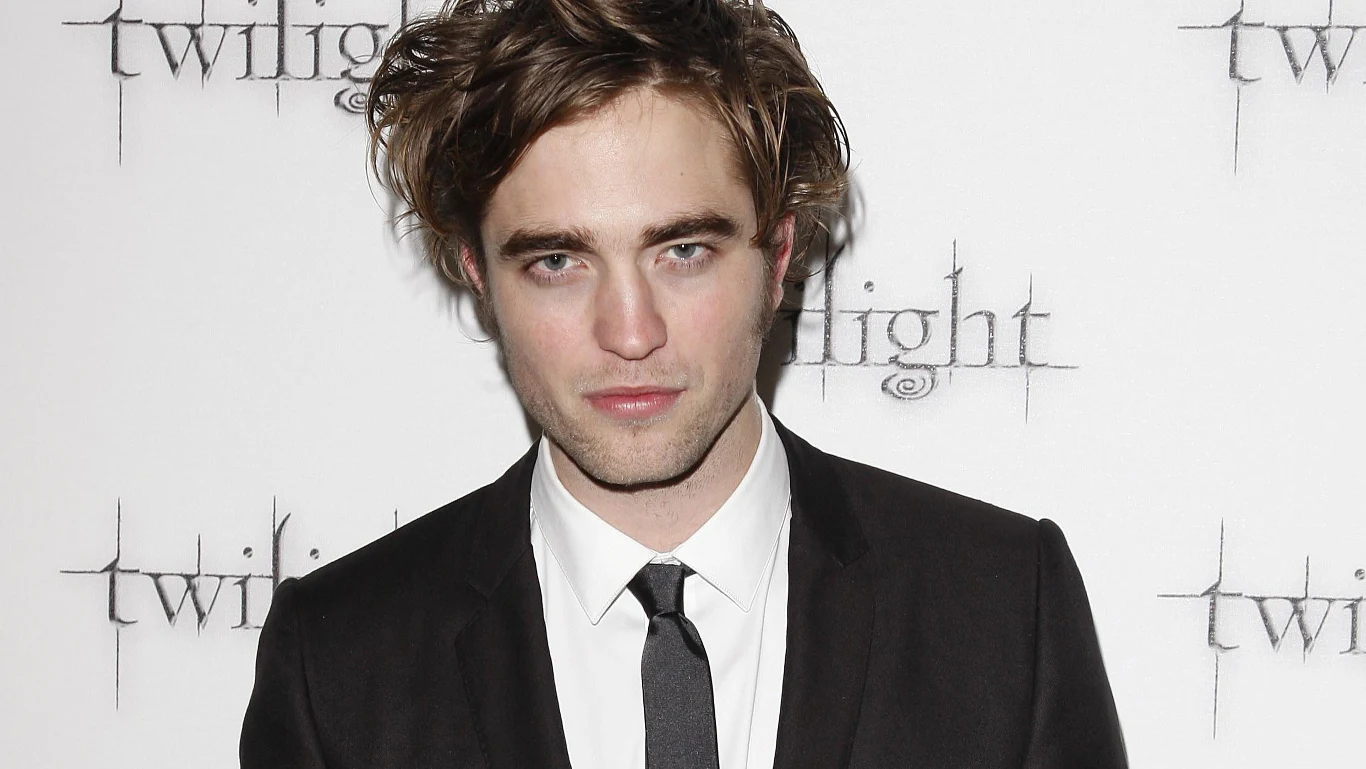 Robert Pattison (2008 r.) Mężczyzna w eleganckim, czarnym garniturze i białej koszuli, z krawatem, na tle ścianki promocyjnej z napisem 'Twilight', z rozczochranymi włosami i lekkim zarostem, patrzący prosto w obiektyw.