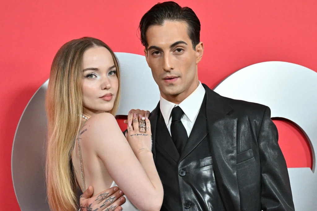 Dove Cameron i Damiano David poznali się w 2023 r. Kobieta o długich, blond włosach obejmuje mężczyznę w czarnym skórzanym garniturze i białej koszuli. Oboje pozują do zdjęcia na tle z dużymi, jasnymi literami i czerwonym tłem.