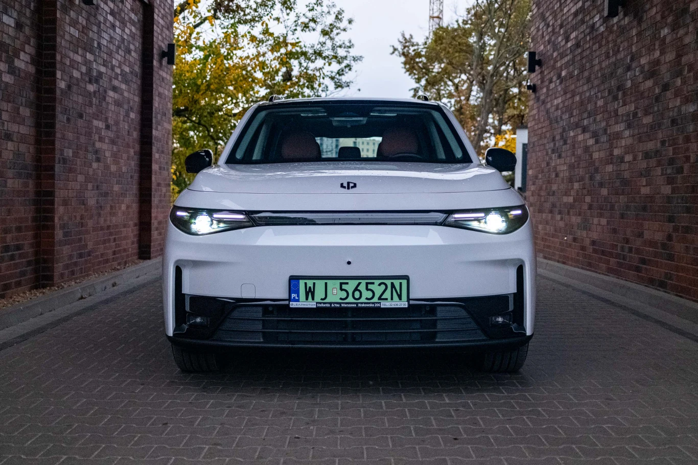 Biały samochód elektryczny z włączonymi światłami, zaparkowany pomiędzy ceglanymi budynkami, na tle drzew z jesienną kolorystyką.