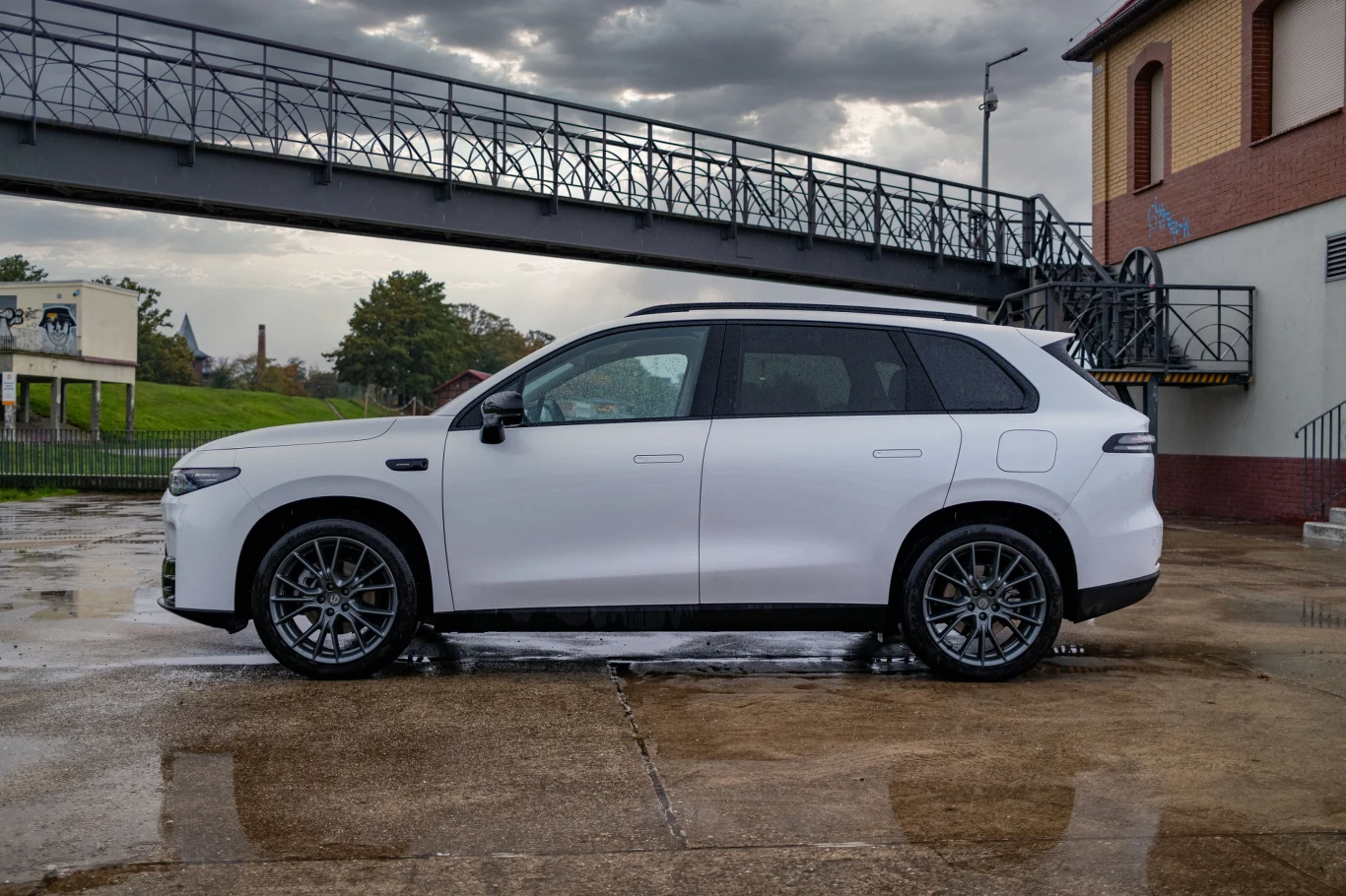 Biały samochód typu SUV zaparkowany na betonowym placu, w tle widoczny budynek z czerwonej cegły oraz metalowa kładka dla pieszych. Na drugim planie drzewa i fragmenty przemysłowej infrastruktury.