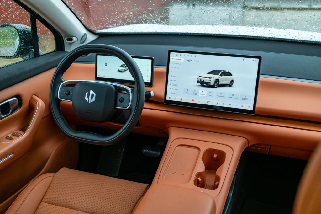 Kokpit samochodu z nowoczesnym, skórzanym wykończeniem w kolorze brązowym, duży dotykowy ekran multimedialny na środku deski rozdzielczej oraz drugi ekran za kierownicą. Wnętrze zaprojektowane minimalistycznie, z wygodnymi miejscami na napoje.