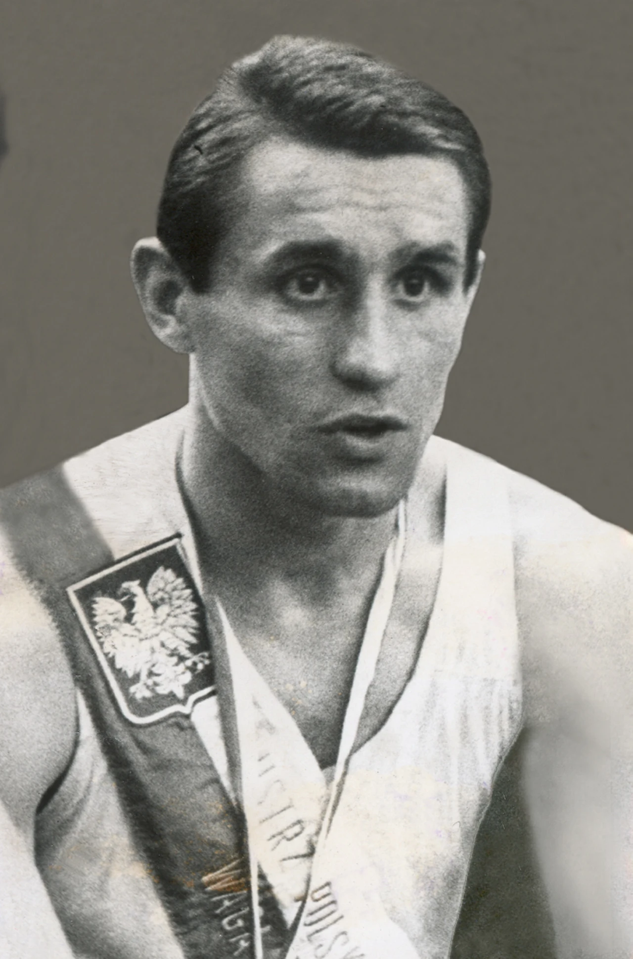 Mężczyzna w sportowym stroju z szarfą dekoracyjną na ramieniu, na której widnieje polski orzeł oraz napisy. Sportowiec ma poważny wyraz twarzy i krótkie ciemne włosy.