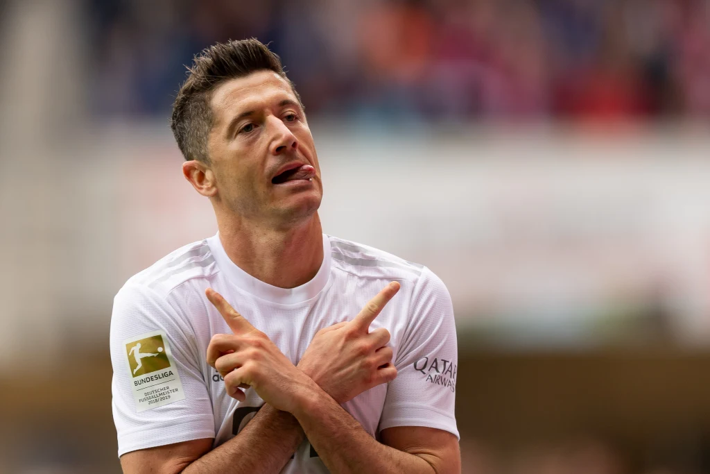 Piłkarz w białej koszulce wykonuje gest z założonymi skrzyżowanymi rękoma, na jego twarzy widać skoncentrowany wyraz. W tle rozmazana publiczność na trybunach.