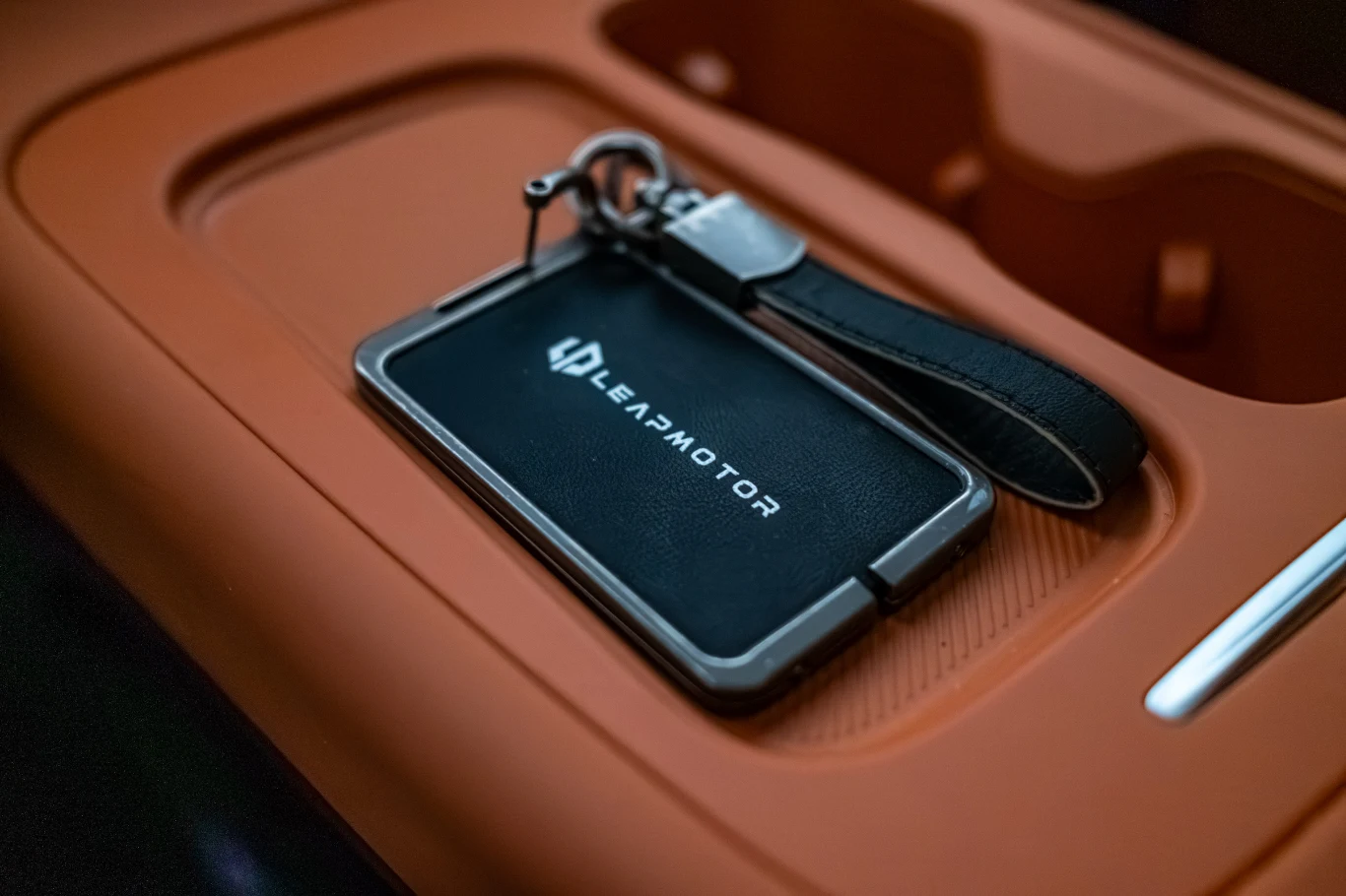Czarny brelok z logiem LEAPMOTOR oraz skórzanym paskiem umieszczony na pomarańczowej powierzchni, prawdopodobnie w schowku wewnątrz samochodu.