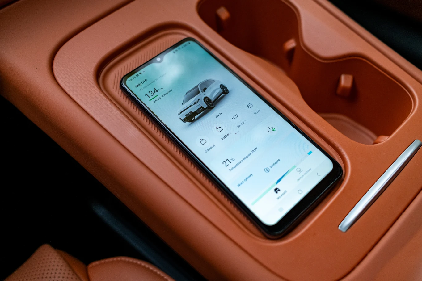 Smartfon umieszczony w konsoli środkowej samochodu, na ekranie wyświetlona aplikacja obsługująca funkcje pojazdu takie jak klimatyzacja, poziom ładowania baterii i lokalizacja auta; wnętrze wykonane z brązowej skóry i plastiku.