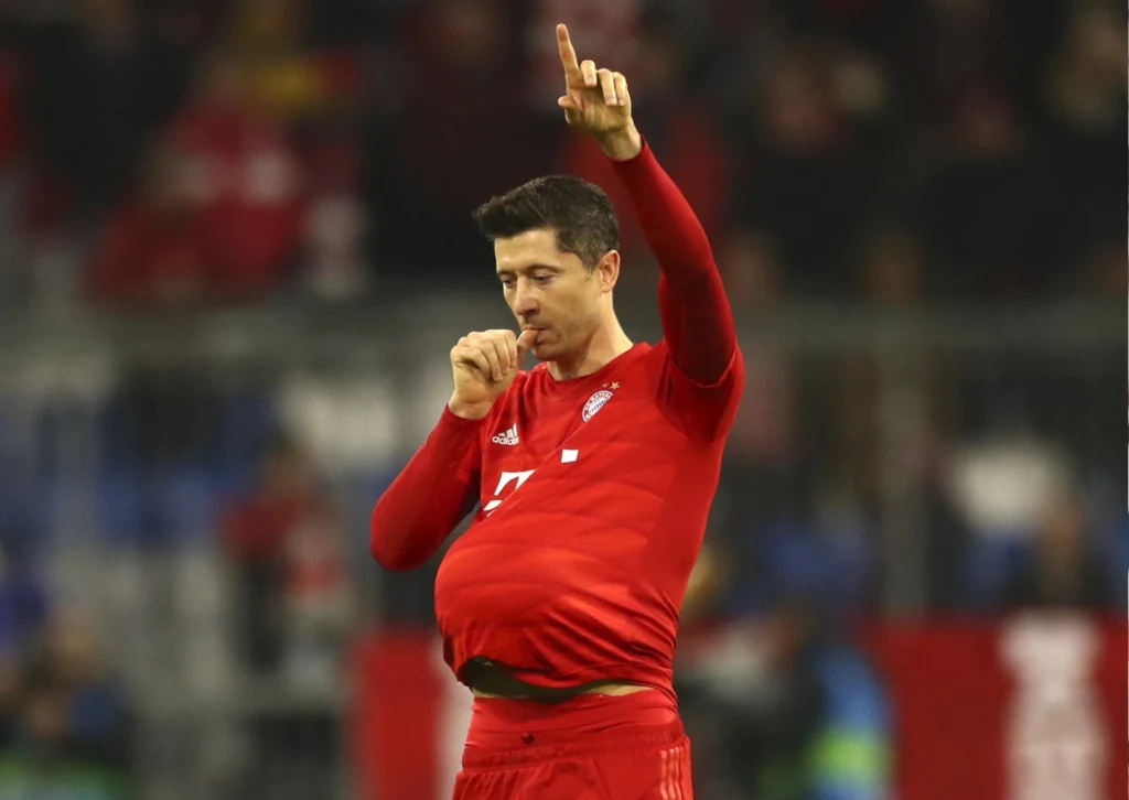 Piłkarz w czerwonym stroju sportowym wykonuje gest ssania kciuka, mając jednocześnie piłkę lub inną wypukłość pod koszulką, co jest symbolicznym gestem nawiązującym do tematów rodzinnych lub radości z okazji narodzin dziecka.