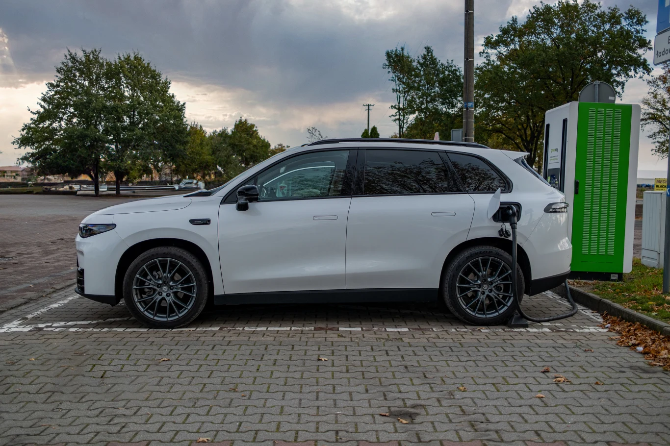Biały samochód elektryczny typu SUV zaparkowany przy stacji ładowania, podłączony do zielonego terminala na tle parkingu otoczonego drzewami. Po bokach widoczne fragmenty zieleni i kostki brukowej.