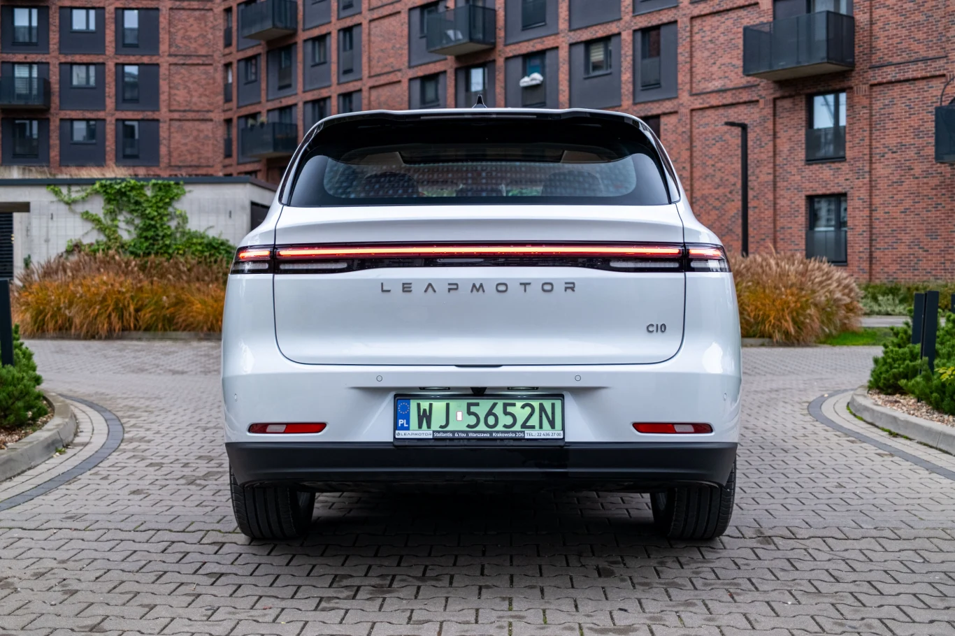 Tył białego samochodu elektrycznego marki Leapmotor C10, zaparkowanego na brukowanej uliczce wśród nowoczesnych budynków z cegły. Na pojeździe widoczna jest zielona tablica rejestracyjna.
