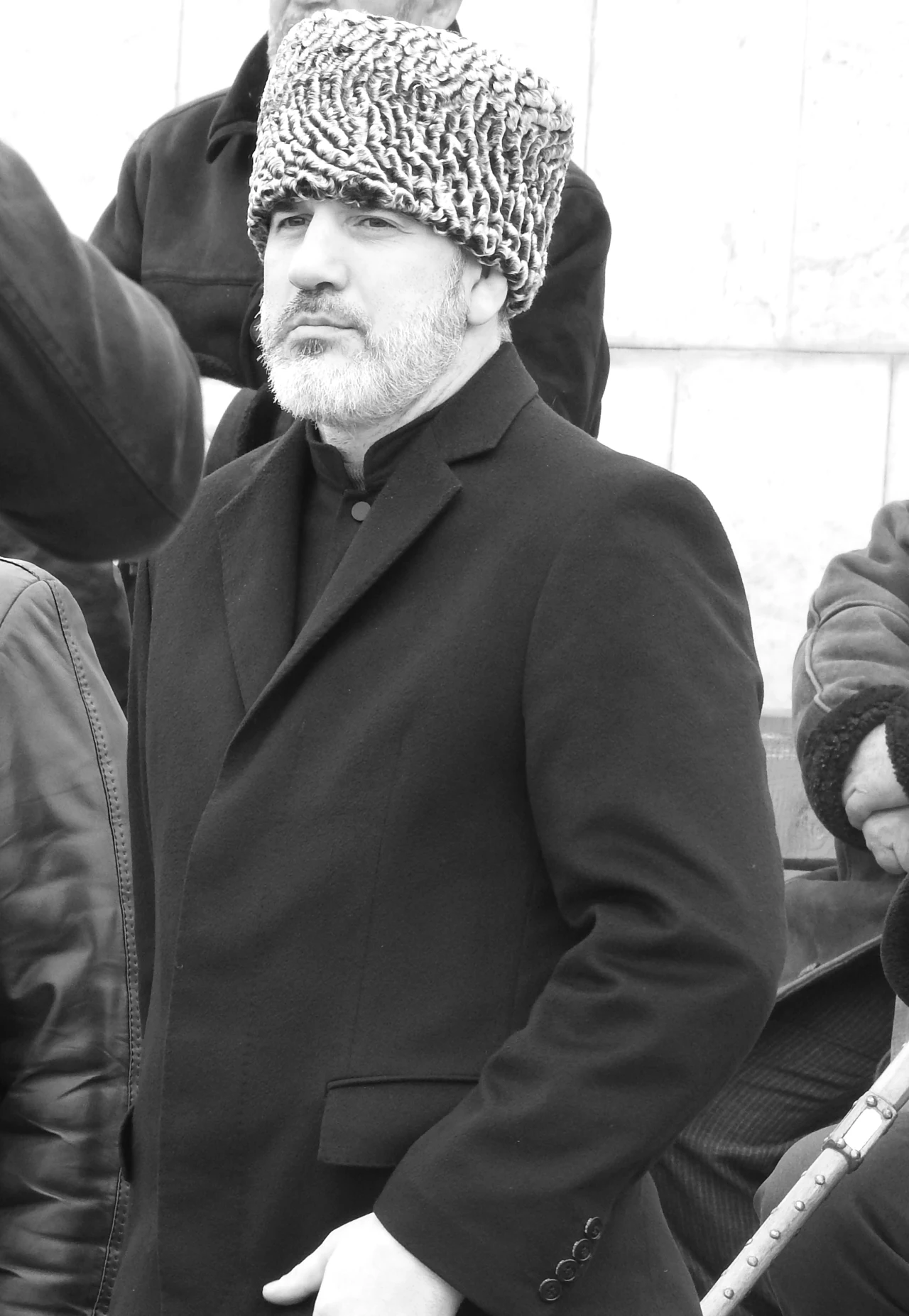 Mężczyzna w czarnym płaszczu i tradycyjnej futrzanej czapce stoi w otoczeniu innych osób.