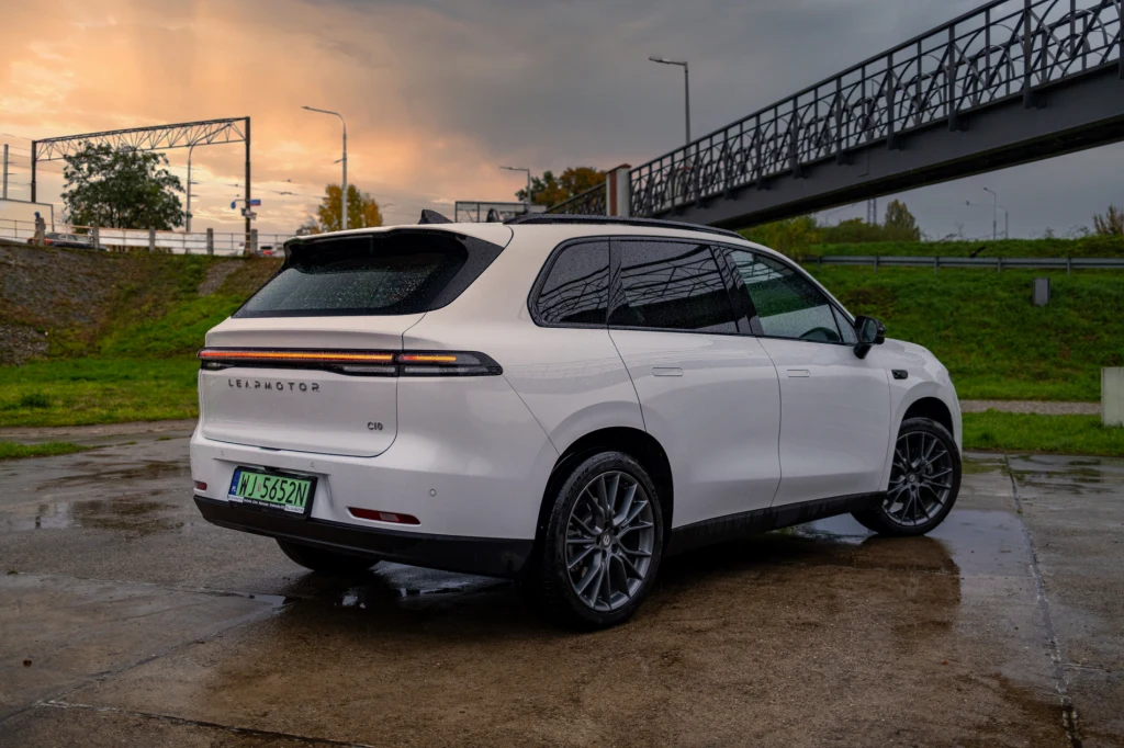 Biały, nowoczesny SUV marki Seres zaparkowany na mokrej nawierzchni, okolica otoczona zielenią i metalową kładką dla pieszych, wilgotna Aura oraz zachmurzone niebo z widocznym zachodem słońca.