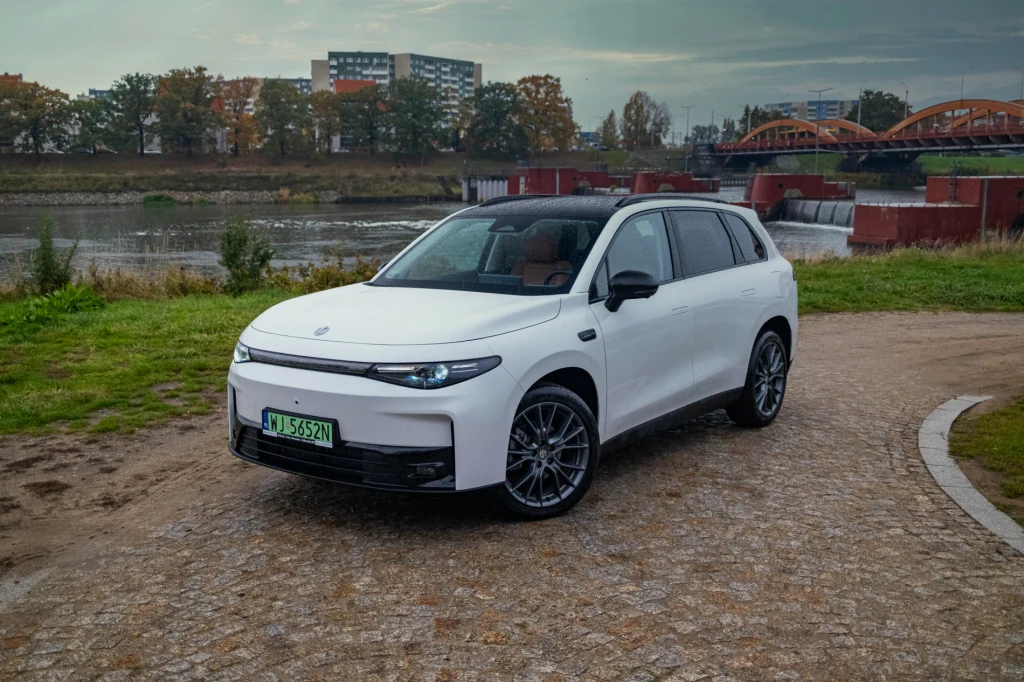 Nowoczesny biały SUV elektryczny stoi na brukowanej alejce nad rzeką, w tle widoczne są zielone drzewa, niska zabudowa oraz most o pomarańczowych łukach.