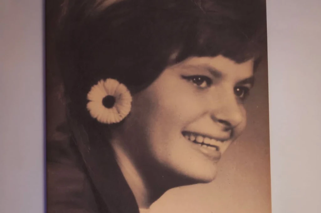 Krysia Uśmiechnięta młoda kobieta w czarno-białym ujęciu, z krótkimi, ciemnymi włosami, nosząca dużą kwiatową ozdobę na uchu, patrząca w bok.