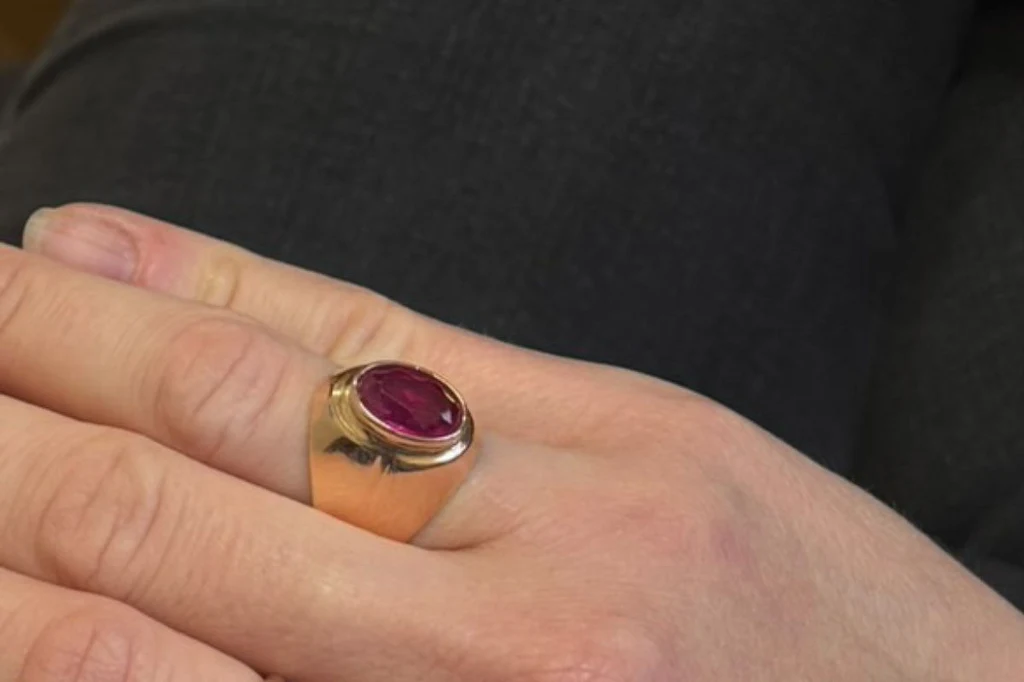 Pierścionek z rubinem Dłoń osoby z dużym złotym pierścieniem z czerwonym kamieniem osadzonym centralnie, widoczne naturalne paznokcie, tło ciemne i jednolite.